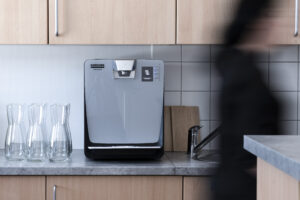 Ein Fotoausschnitt einer Küche, in der sich ein silberner Getränkeautomat sowie Glaskaraffen links davon befinden. Auf der rechten Seite läuft eine schwarz gekleidete Person durch das Bild.