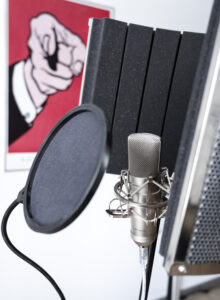 Ein Foto von einem Mikrofon, um das Abdeckungen angebracht sind. Rechts oben hängt ein Bild an der Wand mit einem knallig roten Hintergrund und einem ausgestreckten Zeigefinger darauf.