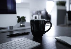 Ein Fotoausschnitt eines Schreibtisches, auf dem sich eine schwarze Tasse, eine weiße Tastatur mit Maus sowie ein schwarzer Kugelschreiber mit der Aufschrift „STORMING“ befinden. In der linken oberen Ecke ist ein Ausschnitt eines Bildschirmes zu sehen.