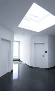 Ein Ausschnitt eines Flurs, welcher Zugang zu drei verschiedenen Türen bietet. An der Decke scheint aus einem viereckigen Dachfenster helles Licht herein. Die Wände sind weiß gestrichen und der Steinboden ist dunkelgrau.